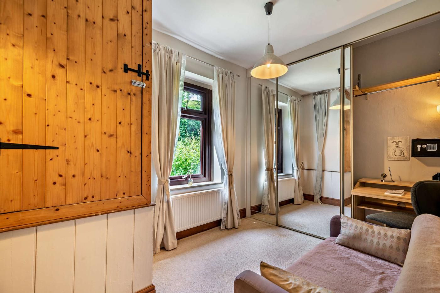 A room with a wooden wardrobe, a window with curtains, a mirror, a desk with a lamp, and a bed with pillows.