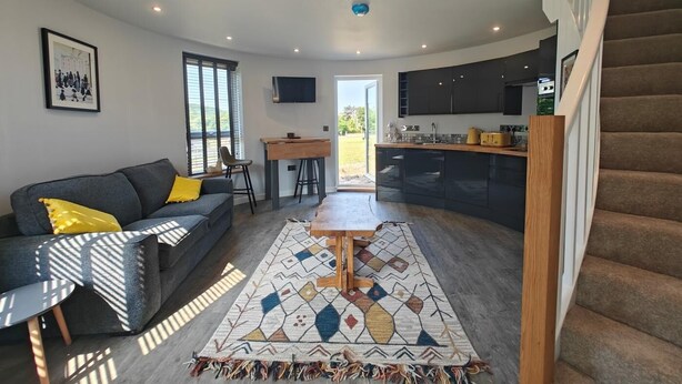 Um espaço de estar moderno com sofá, mesa de centro e tapete estampado. É visível uma área de cozinha com armários escuros e bancada de madeira.