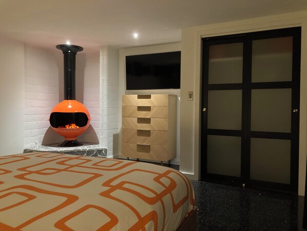 Una habitación con chimenea moderna, cama con ropa de cama estampada, un armario de madera y una puerta con paneles de vidrio esmerilado.