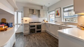 Une cuisine moderne avec des meubles blancs, une cuisinière à gaz, deux fours et une fenêtre donnant sur une façade en pierre.