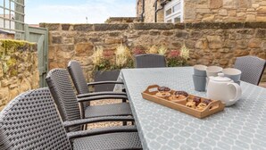 Une terrasse avec des fauteuils en osier et une table préparée pour le thé, entourée de murs en pierre et de plantes.