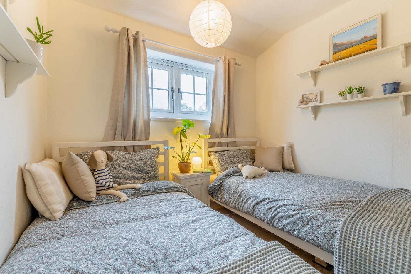 A bedroom with two beds, a window with curtains, a hanging light fixture, and a shelf with plants.