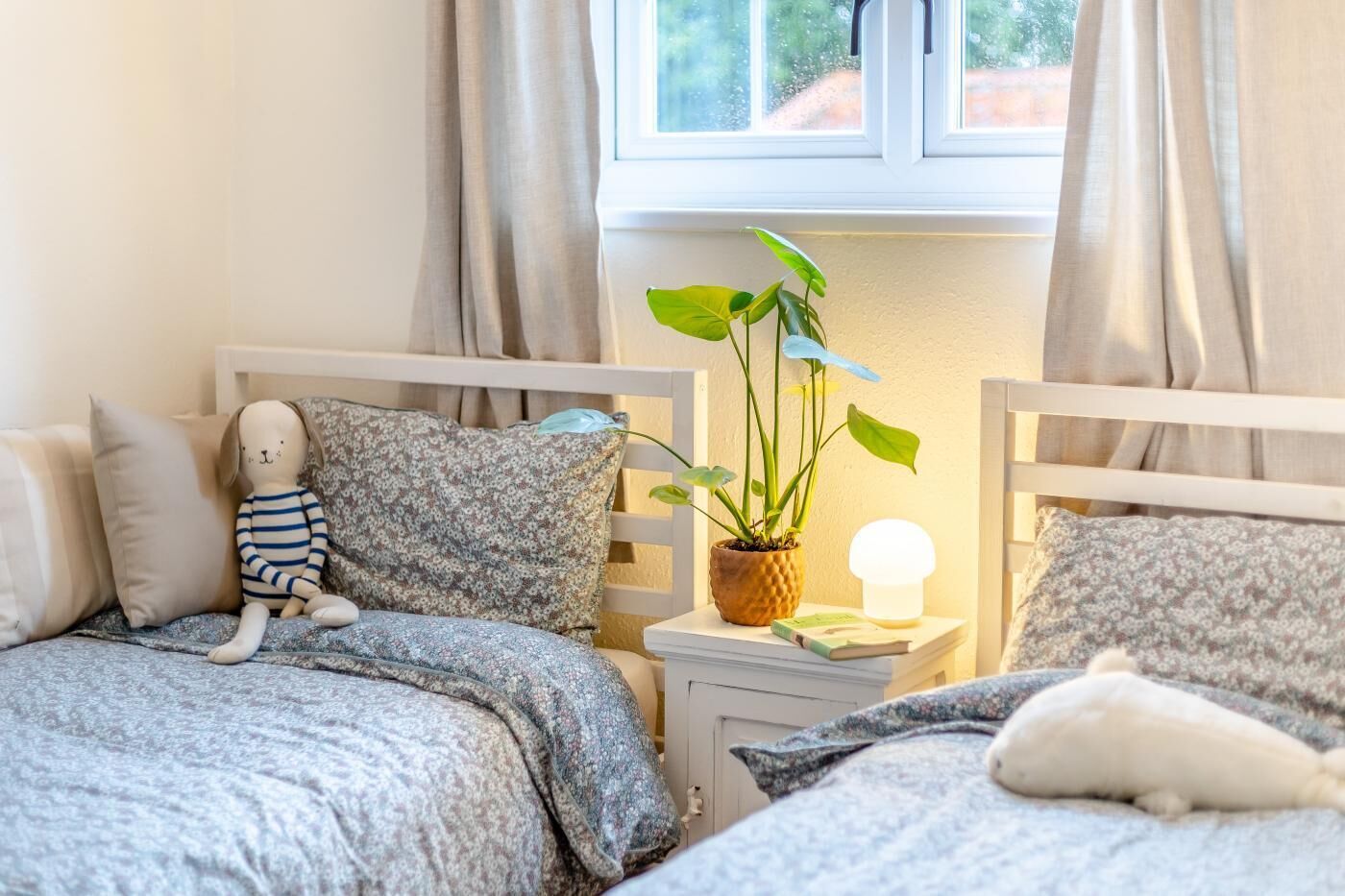 Une chambre confortable avec deux lits, une table de chevet avec une lampe, une plante en pot et un jouet en peluche.