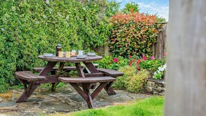 Une table de pique-nique en bois, avec quatre bancs, entourée d’une végétation luxuriante et de plantes en fleurs.