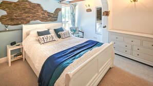A bedroom with a stone accent wall, a white bed with blue bedding, a nightstand, a dresser, and a mirror. - 3 Bed in St. Ervan (oc-t29660) (Saint Ervan)