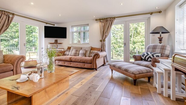 Amplia sala de estar con sofá, reposapiés y mesa de centro, televisión y ventanas grandes con cortinas.