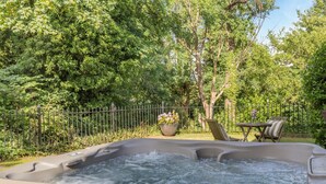 Un jacuzzi rodeado por un exuberante jardín con una valla, una mesa con sillas y una planta en maceta.