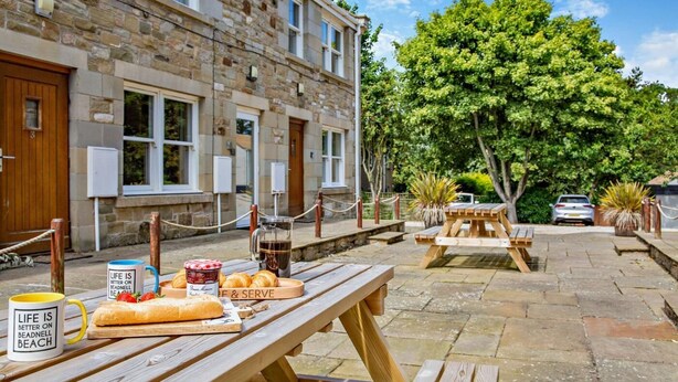 A stone building with wooden doors and windows, outdoor wooden benches, picnic tables, and a tree in the background. - 1 Bed in Beadnell (oc-l29758) (Beadnell)