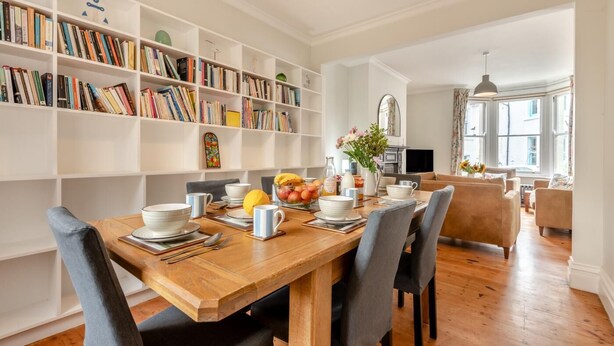 A dining area with a wooden table set for breakfast, surrounded by bookshelves filled with books. - 4 Bed in Swanage (oc-a29454) (Swanage)