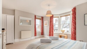 A bedroom with a large bed, a bedside table, a mirror, a radiator, and a window with red curtains. - 4 Bed in Swanage (oc-a29454) (Swanage)