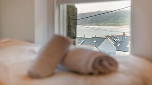 A bed with a rolled towel and a window overlooking a coastal view with buildings. - 3 Bed in Sennen Cove (oc-c29670) (Sennen)