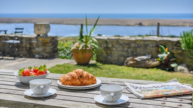 A wooden table with a croissant, strawberries, and cups, overlooking a beach with a stone wall and potted plants. - 2 Bed in Langton Herring (oc-r29848) (Langton Herring)