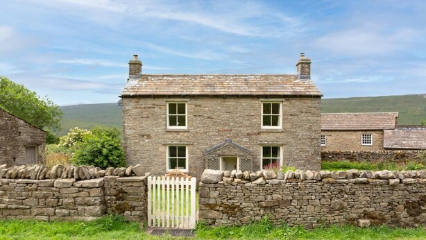 Una casa de piedra con un portón blanco, un porche pequeño y un muro de piedra en un entorno rural.
