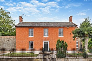 Une maison à deux étages, construite en briques, avec une façade symétrique, une porte centrale et un petit jardin à l’avant.