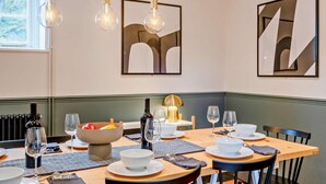 A dining area with a table set for four, including wine glasses, bowls, and plates. There are abstract paintings on the walls and a window with a view of greenery.