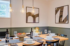 Un coin repas avec une table dressée pour quatre personnes, comprenant des verres à vin, des bols et des assiettes. Les murs sont ornés de peintures abstraites et une fenêtre offre une vue sur la verdure.