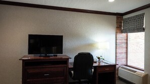 A hotel room with a wooden desk, a television, a chair, a lamp, and a window with blinds. - Executive King Room with with Modern Amenities & Breakfast (Hillsdale)