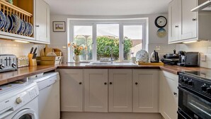Une cuisine avec des meubles blancs, un plan de travail en bois, un lave-linge, une cuisinière et une fenêtre donnant sur un paysage verdoyant.