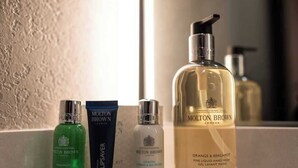 Ein Badezimmerschrank mit verschiedenen Pflegeprodukten, darunter eine grüne Flasche, eine blaue Tube, eine klare Flasche und eine große Flasche Handseife mit Orange und Bergamotte.