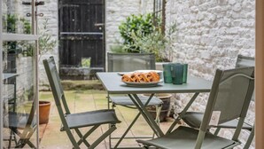 A small outdoor dining area with a table and chairs, a plate of food, and a window with a view of a stone wall and greenery. - 3 Bed in Swanage (oc-n29755) (Swanage)