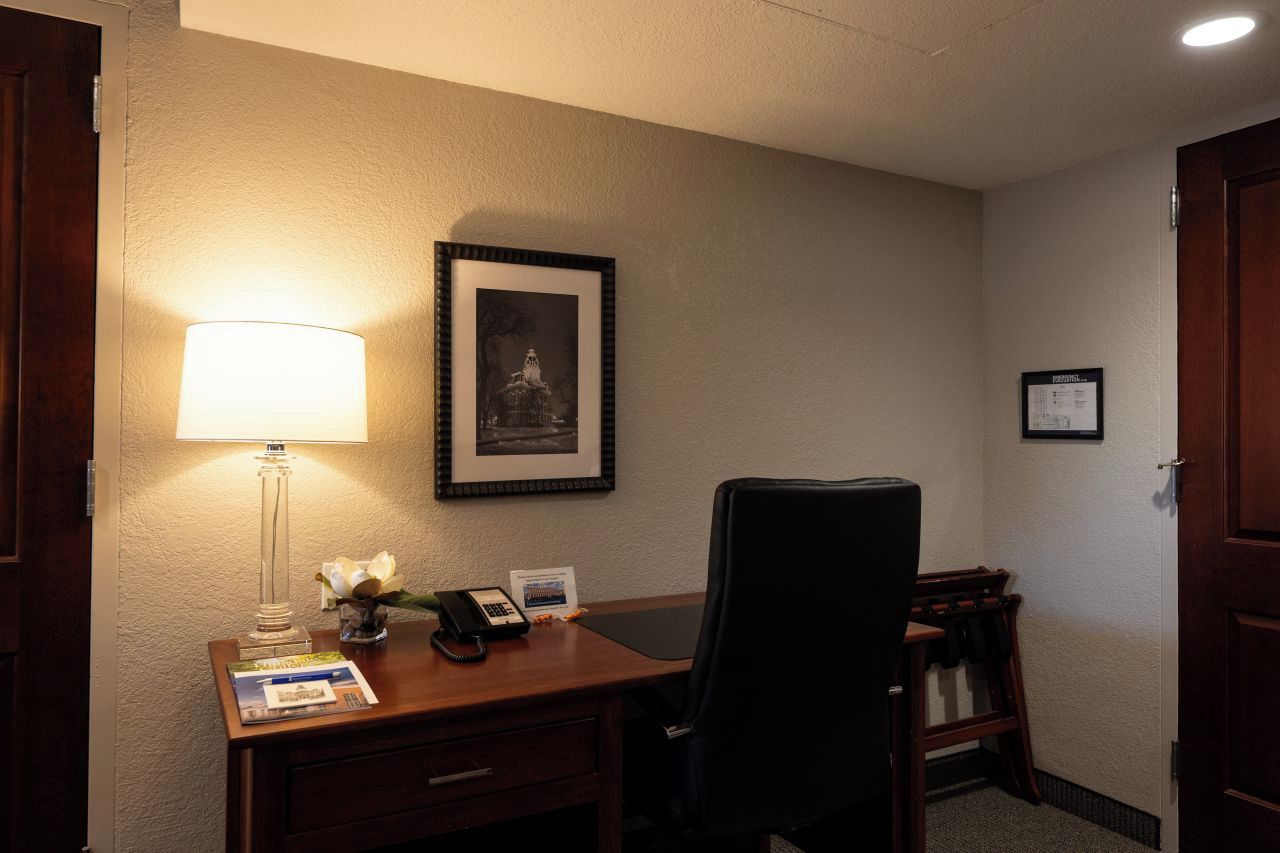 Una habitación de hotel con un escritorio, una lámpara, un cuadro enmarcado, un teléfono y una silla.