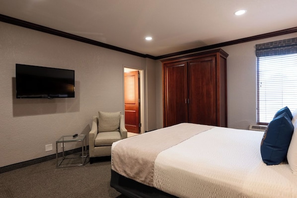 A hotel room with a bed, a TV mounted on the wall, a chair, and a wardrobe. - Executive King Suite with Sleeper Sofa. Breakfast Included (Hillsdale)