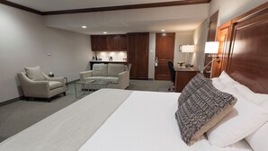 Hotelkamer met een groot bed, twee fauteuils, een kleine tafel, een bureau, een lamp en een kitchenette op de achtergrond.