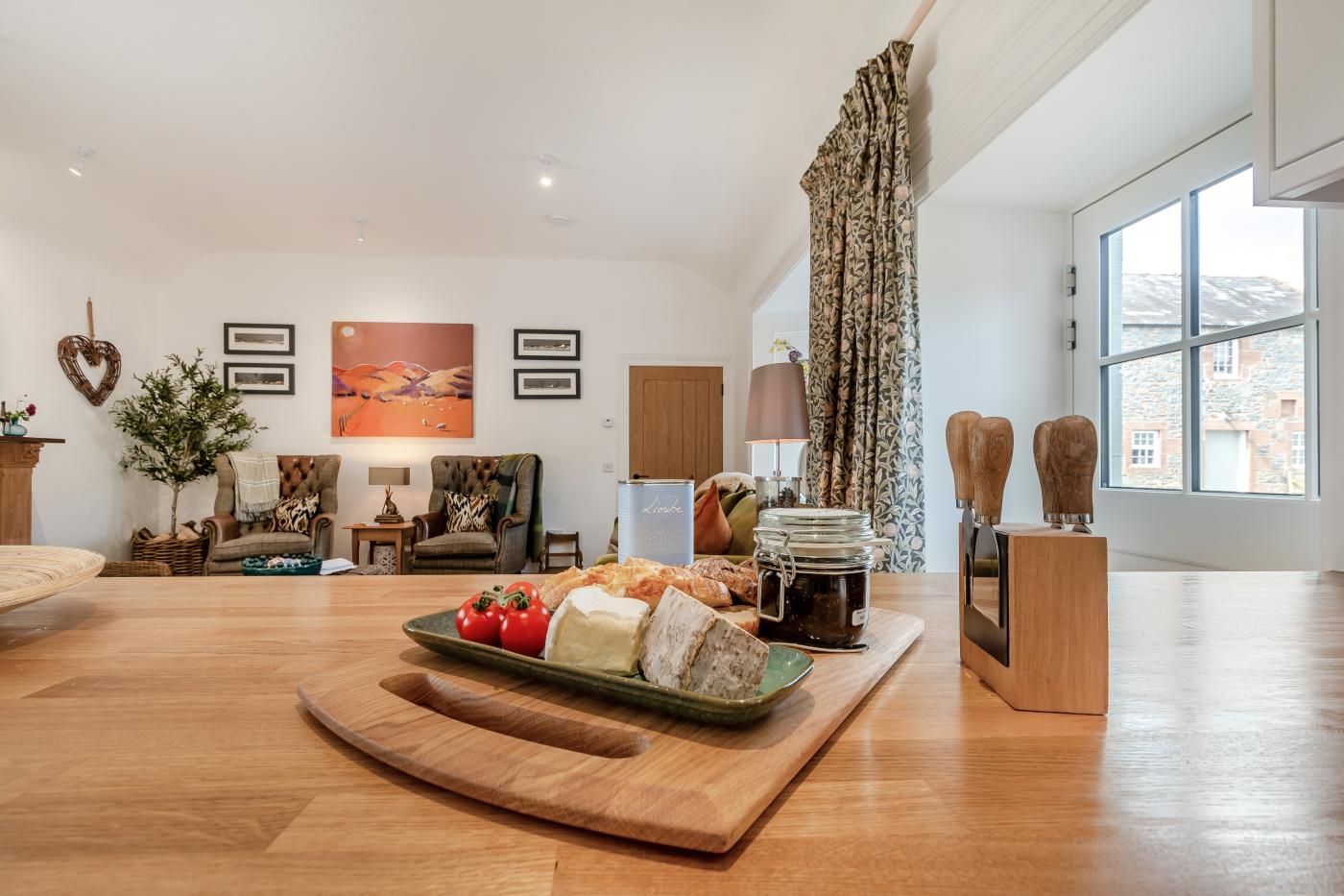 A spacious living room with a wooden floor, a dining table with food, a sofa, a chair, a lamp, and a large window.
