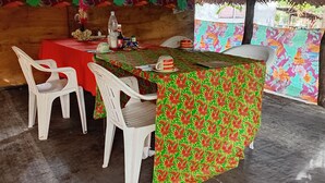 A dining area with a table covered in a red tablecloth, white plastic chairs, and a patterned tablecloth.