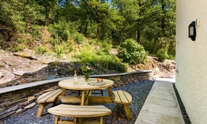 An outdoor wooden table and benches with a stone wall backdrop, a bottle, and a vase with flowers. - 3 Bed in Newby Bridge (oc-c29820) (Newby Bridge)