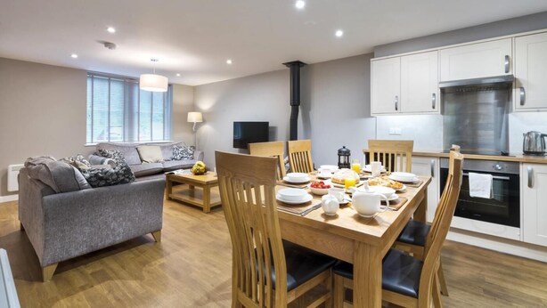 Una sala moderna con un sofá gris, una mesa de comedor de madera y sillas. Un televisor está montado en la pared, y se ve una cocina con armarios y electrodomésticos blancos.
