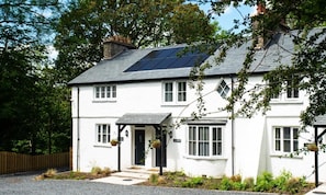 A white two-story house with a black roof, solar panels, and a small porch with hanging baskets. - 2 Bed in Newby Bridge (oc-b29819) (Newby Bridge)