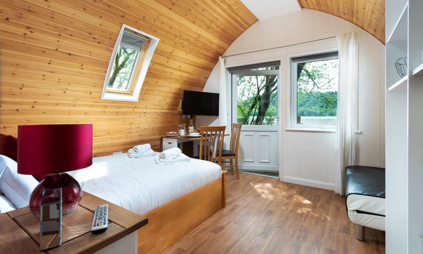 Een slaapkamer met een houten plafond, een bed met witte lakens, een rode lamp, een televisie en uitzicht op groen buiten.