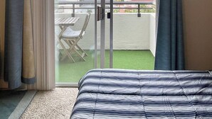 Quarto de hotel com uma cama, porta de vidro deslizante para uma varanda com mesa e cadeiras, e vista para a paisagem urbana.
