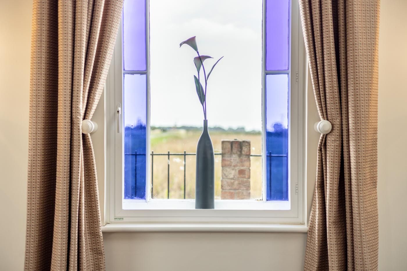 A window with curtains, a vase with a plant, and a view of a brick wall and fence.