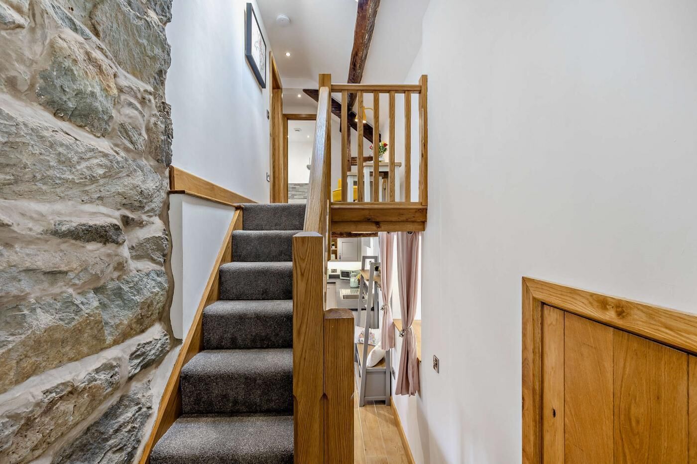 A staircase with stone walls, wooden railings, and carpeted steps leading to a room with a kitchen in the background.