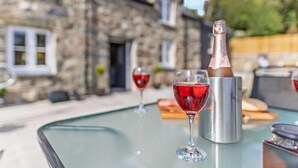 A table with a bottle of champagne and two glasses of red wine, set outdoors with a stone building in the background. - 3 Bed in Brithdir (oc-b29844) (Brithdir and Llanfachreth)