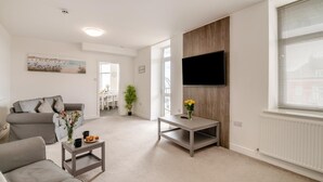 Ein modernes Wohnzimmer mit einem grauen Sofa, einem Couchtisch, einem an der Wand befestigten Fernseher und einem Essbereich im Hintergrund.