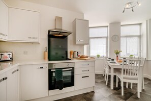 Una cocina moderna con armarios blancos, horno empotrado, tostadora y comedor con mesa y sillas blancas.