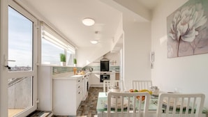 A modern kitchen with white cabinets, a dining area with a table set for two, and a large window with a view of the city. - 2 Bed in Bridlington (oc-o29897) (Bridlington)