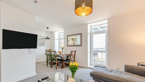 Een moderne woonkamer met een grijze bank, een eethoek met houten stoelen, een aan de muur bevestigde televisie en een groot raam dat natuurlijk licht binnenlaat.