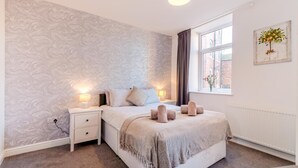 Een slaapkamer met behang met een patroon, een bed met een beige deken, twee nachtlampjes, een raam met uitzicht en een schilderij aan de muur.
