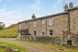 A stone building with multiple windows and a small door, surrounded by a grassy area and a stone wall. - 3 Bed in Gunnerside (oc-e26693) (Ivelet)