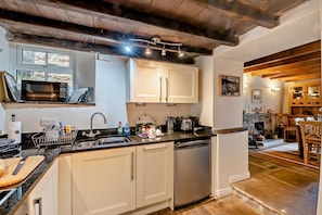 A kitchen with white cabinets, a dishwasher, and a microwave. There is a dining area with wooden furniture and a fireplace. - 3 Bed in Gunnerside (oc-e26693) (Ivelet)