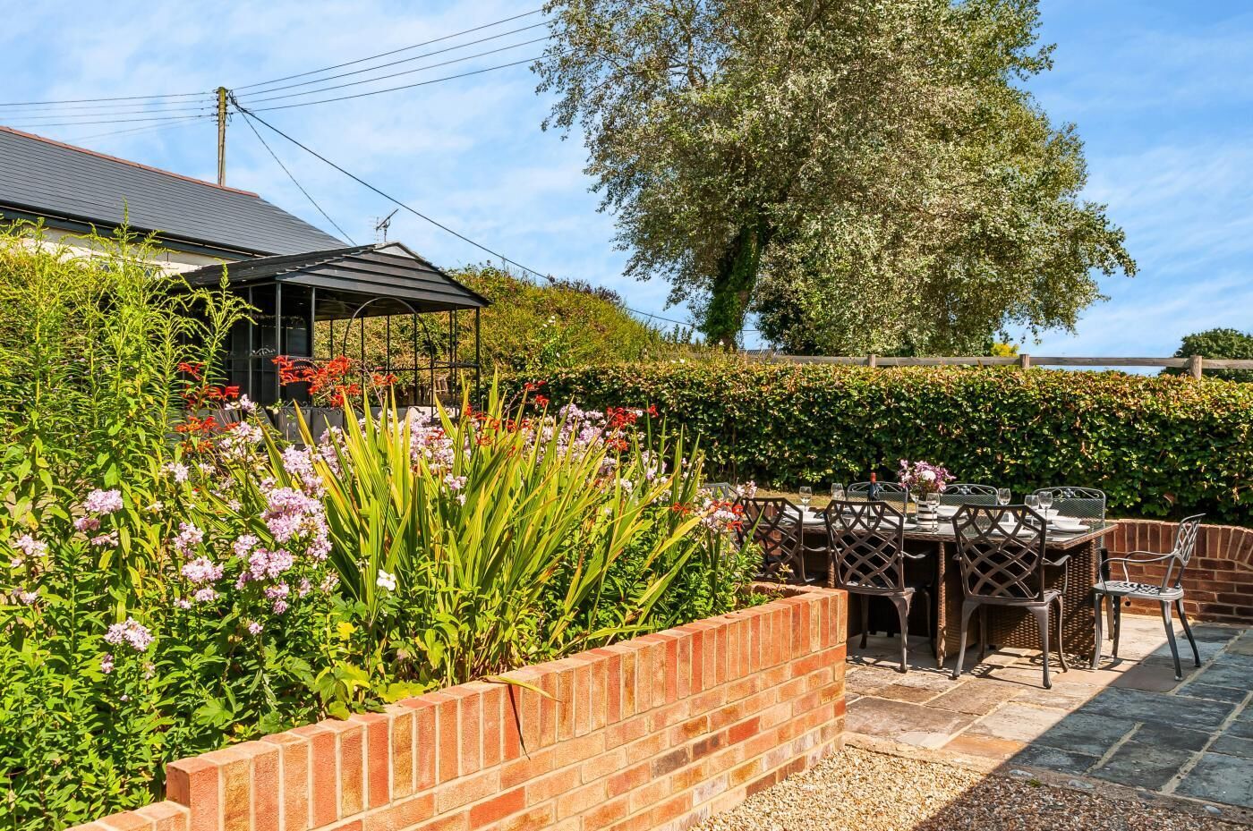 A patio area with a brick wall, outdoor table and chairs, and a variety of flowering plants.