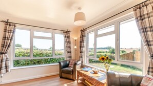 A room with a wooden floor, a leather sofa, a wooden table with a bowl and books, and large windows with curtains. - 4 Bed in Woolage Green (oc-f29907) (Woolage Green)