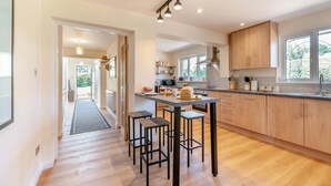 A modern kitchen with wooden cabinets, a central island with stools, and a view of the outdoors through large windows. - 4 Bed in Woolage Green (oc-f29907) (Woolage Green)