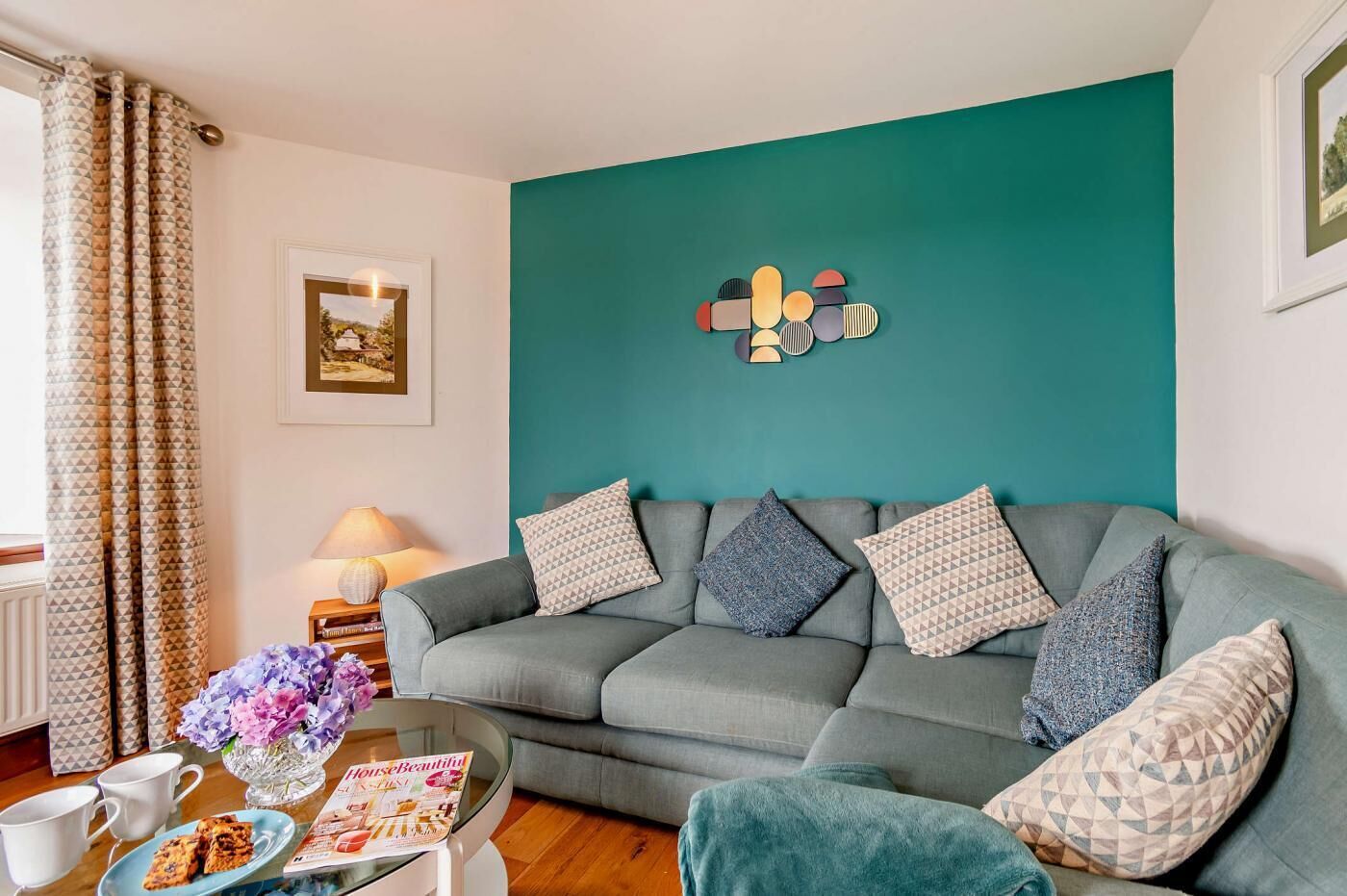 Een woonkamer met een muur in turquoise, een grijze bank met gekleurde kussens, een bijzettafeltje met een tijdschrift en een vaas met bloemen, en een raam met behang.