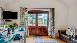 Un comedor con una mesa de madera dispuesta para cuatro personas, un televisor montado en la pared y vistas a través de una puerta de madera grande.
