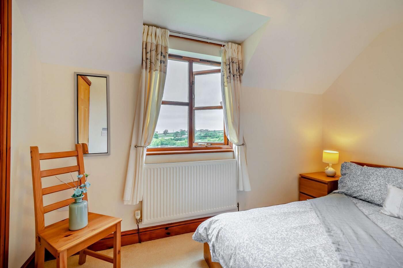 Une chambre à coucher avec un lit, une table de chevet avec une lampe, une chaise avec un vase, une fenêtre avec des rideaux et un radiateur.
